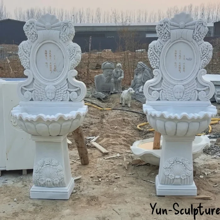 A pair of ornate holy water wall fonts hand-carved from single blocks of natural white marble. Featuring detailed cherub reliefs and grape clusters symbolizing the Eucharist, these stoups are designed to hang securely at church entrances, providing a durable and sacred vessel for liturgical use.