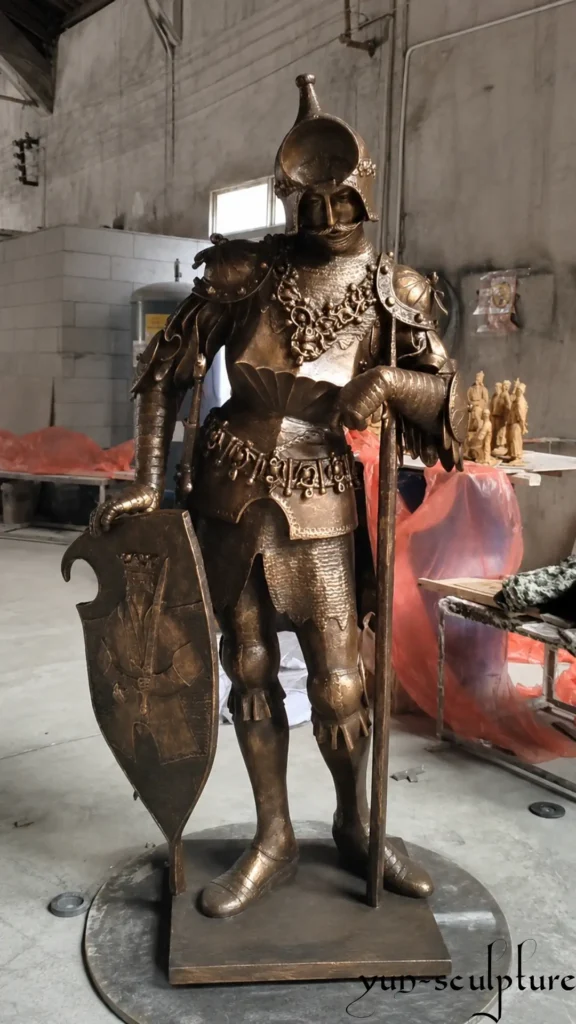 A life-size cast bronze knight statue in full Gothic plate armor at the Yun Sculpture foundry. The design emphasizes "visual mass" through wide pauldrons and an elevated helmet, creating an imposing presence of armored authority. The vertical sword position ensures maximum structural integrity and minimal leverage stress for permanent outdoor installation.