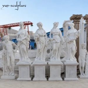 A collection of four life-size hand-carved white marble Greek goddess statues mounted on high pedestals at the Yun-Sculpture outdoor workshop. These elegant figurative sculptures illustrate how classical deities can serve as architectural landmarks for high-end garden landscapes and sunlit terraces.