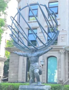 A monumental cast bronze statue of the Titan Atlas supporting a large metal-ringed celestial sphere, set against a grand stone building. Following the accurate Greek mythological tradition of holding up the sky rather than a physical globe, this heroic-scale sculpture by Yun-Sculpture serves as a powerful architectural landmark for corporate plazas and large estates.
