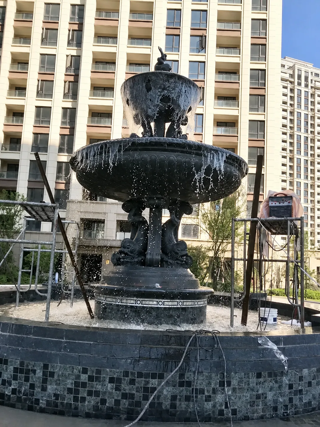 Dark bronze two-bowl fountain running water during a site test with temporary equipment visible