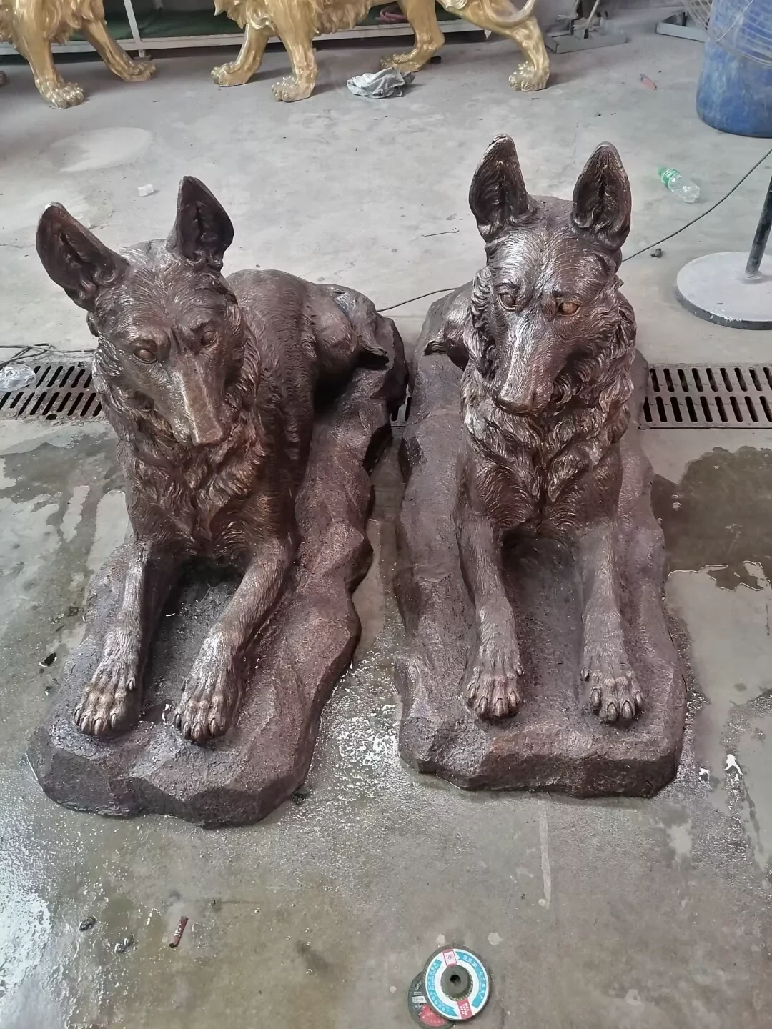 Factory photograph of two reclining bronze German Shepherd sculptures on separate shaped bases.