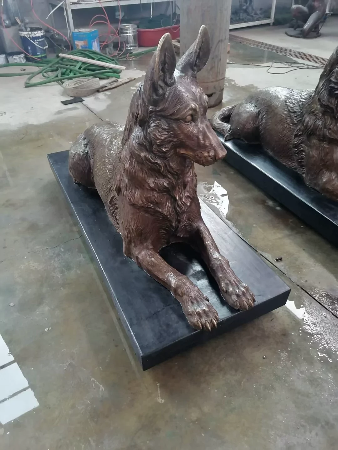 Factory photograph of a reclining bronze German Shepherd sculpture with upright ears, extended front paws and a dark display platform.