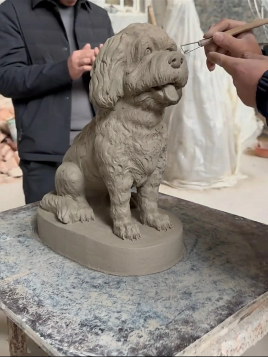 Hands using modelling tools to refine the face of a seated curly-coated clay dog model on a round platform.