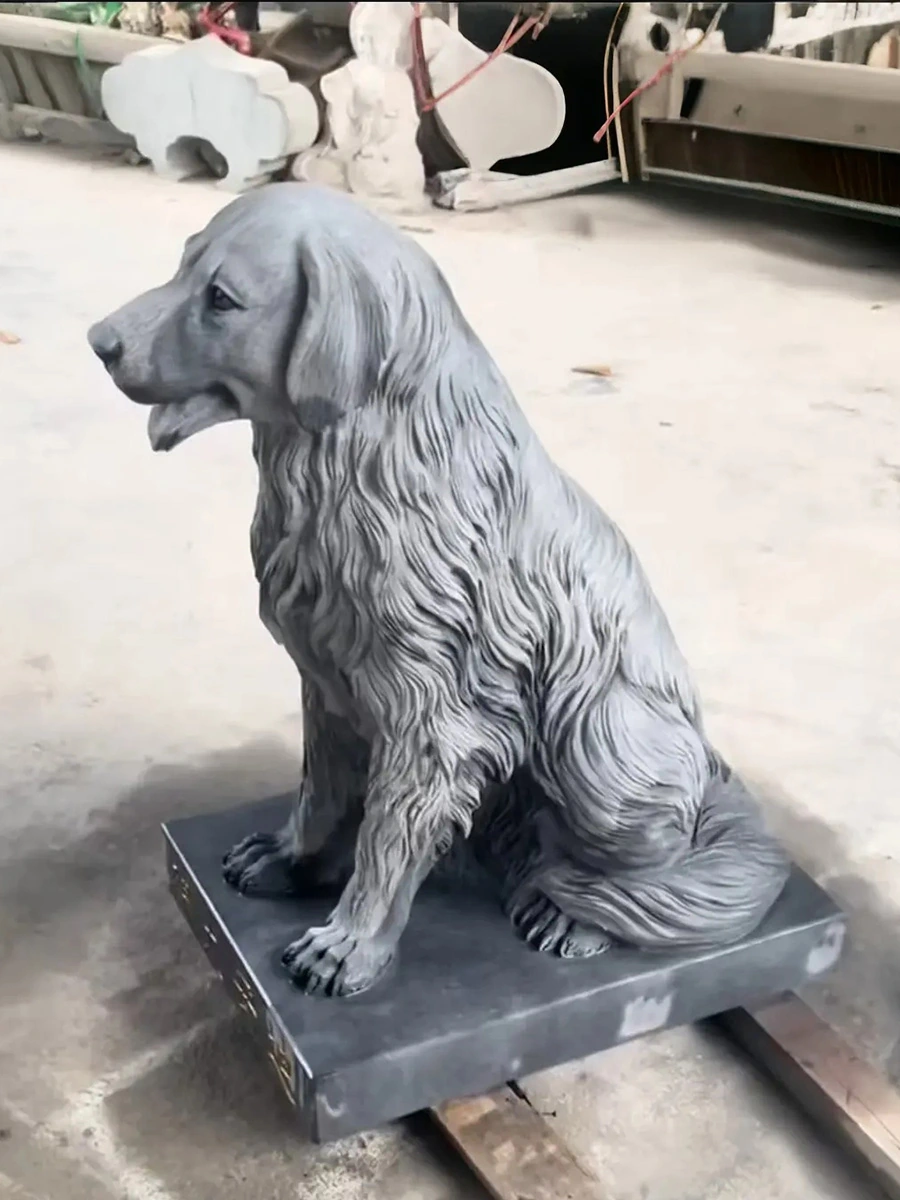Three-quarter view of a grey marble seated long-coated dog sculpture with a curved tail and rectangular base in the workshop.