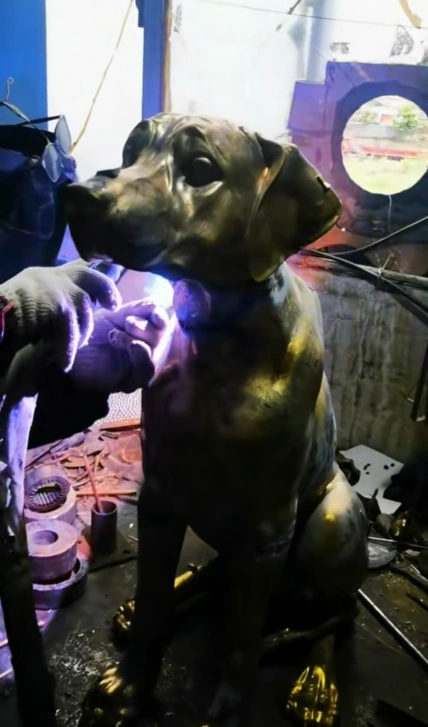 Worker using a tool at the neck of a seated metal dog sculpture, with a bright work area visible in the workshop.