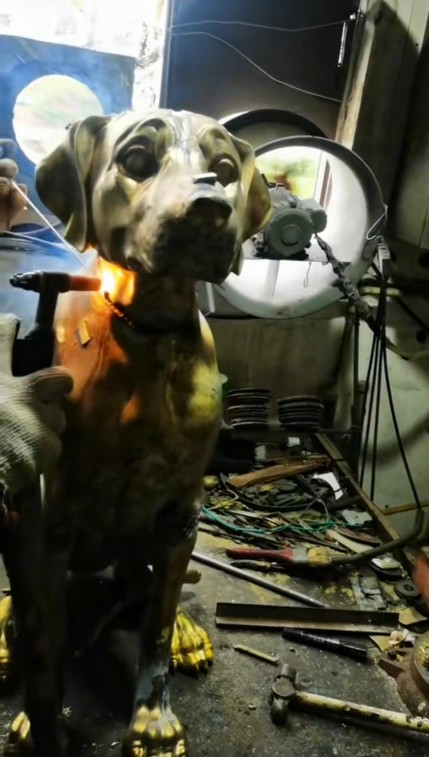 Seated metal dog sculpture with a worker tool and a bright joining area visible near the neck in the workshop.