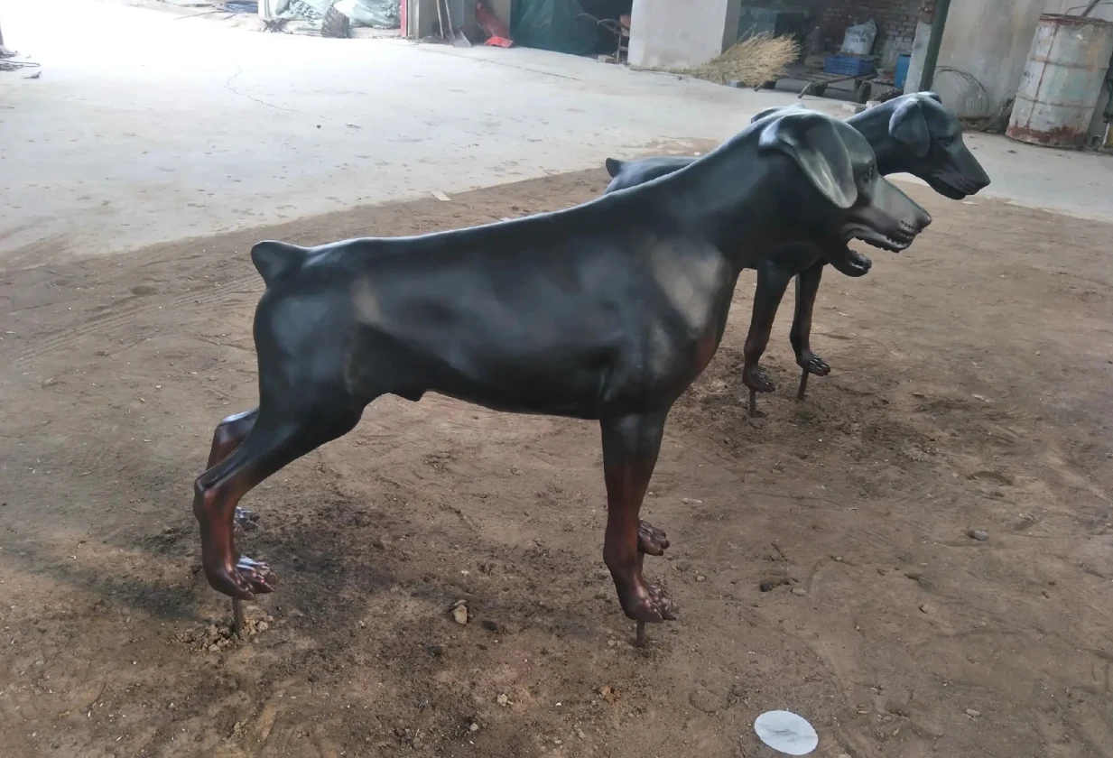 Side view of a dark-finished standing metal dog sculpture showing its head, legs and tail on the workshop floor.