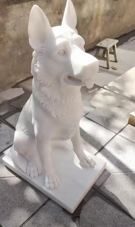 White marble seated dog sculpture with upright ears, a turned head and a rectangular base photographed on paving.